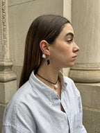A woman in profile wearing a light blue and white striped button-down shirt, a black cord necklace with a white flower pendant, and silver-toned earrings with black dangling charms, set against a stone architectural background.