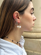 A close-up side profile of a woman wearing a silver hoop earring with dangling clear crystal and pearl charms, along with a beaded choker necklace, against a neutral stone wall background.