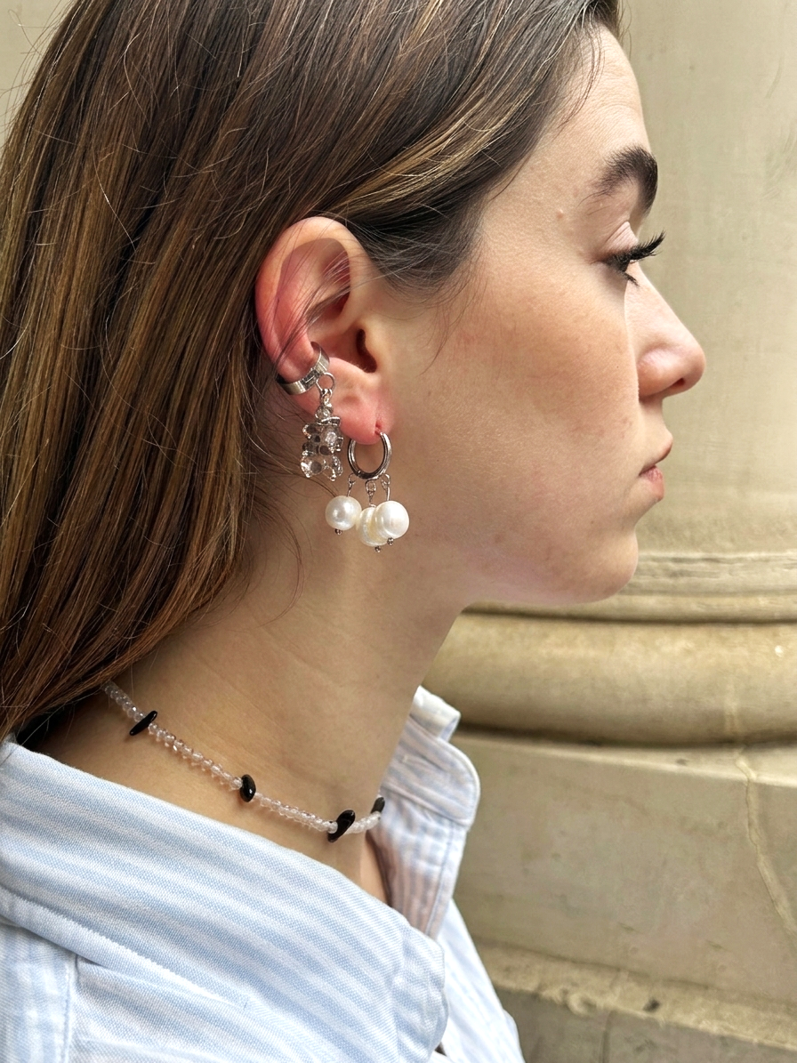 A close-up side profile of a woman wearing a silver hoop earring with dangling clear crystal and pearl charms, along with a beaded choker necklace, against a neutral stone wall background.