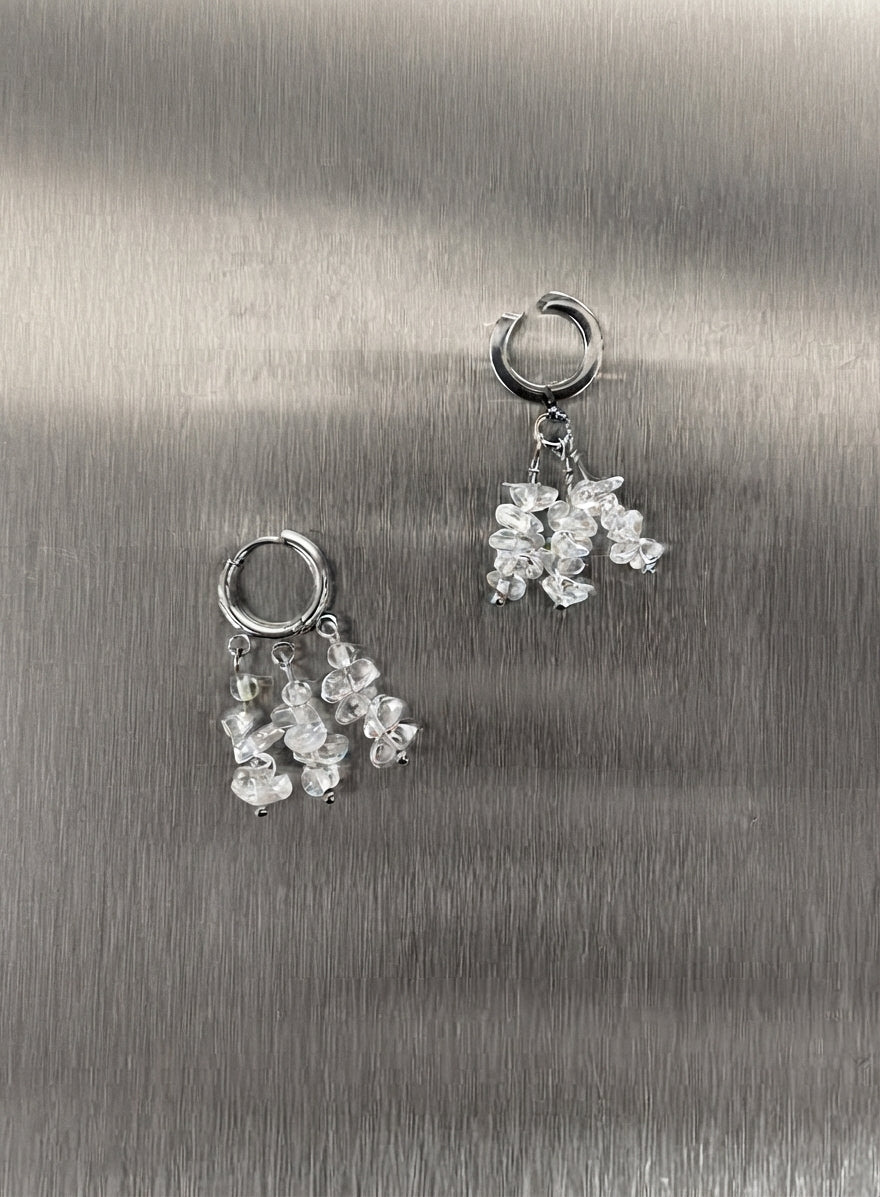 A pair of silver hoop earrings with dangling clusters of clear, faceted crystal-like beads, displayed on a brushed metallic silver surface.