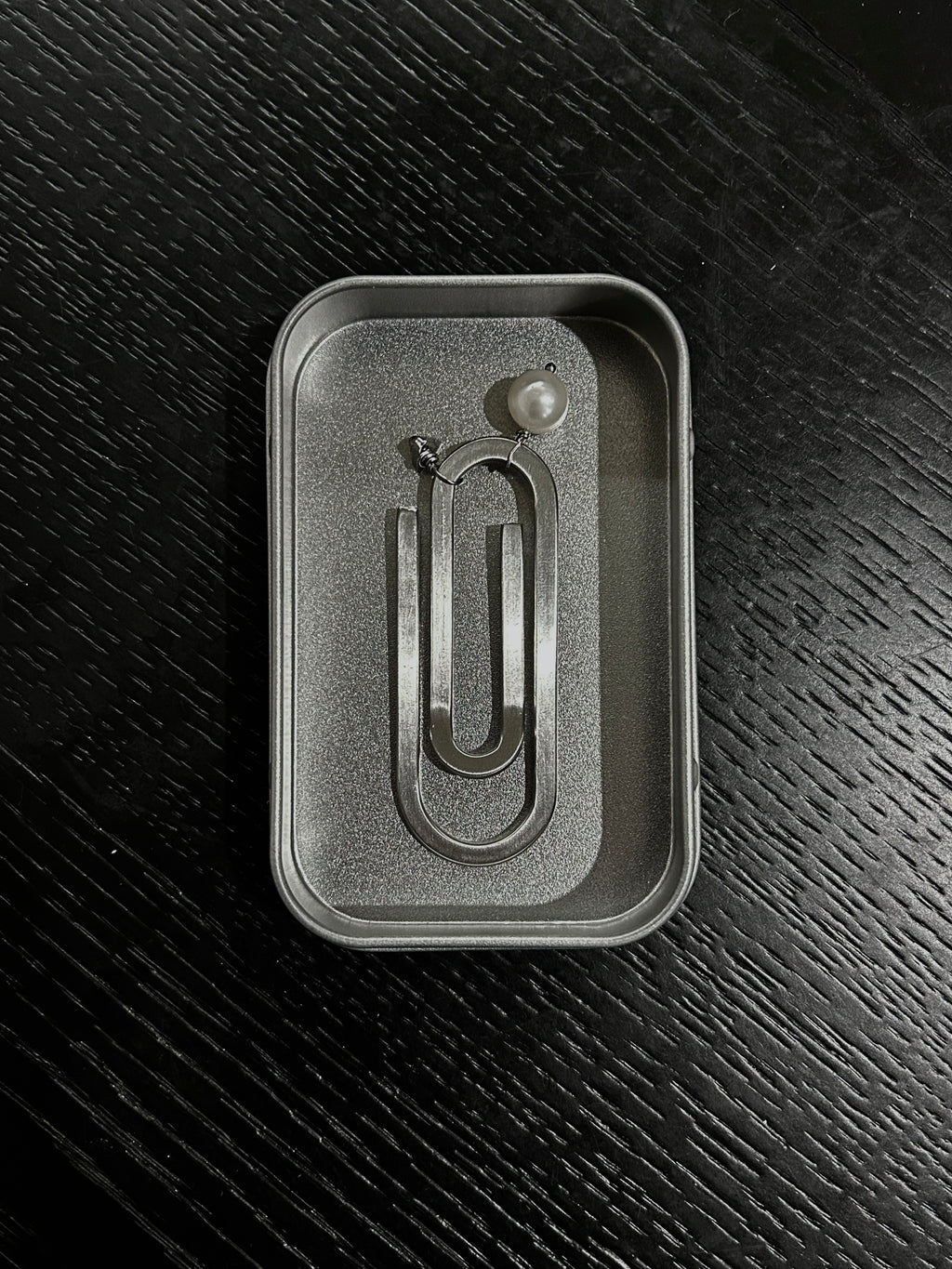 A silver metal paperclip-shaped bookmark with a cultured pearl and initial charm displayed inside its textured steel box insert on a dark wood background.