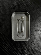 A silver metal paperclip-shaped bookmark with a cultured pearl and initial charm displayed inside its textured steel box insert on a dark wood background.