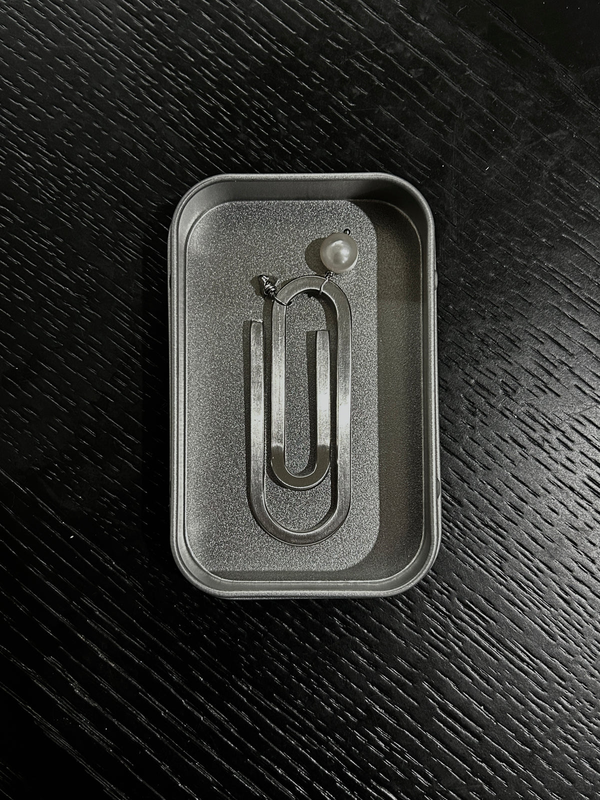 A silver metal paperclip-shaped bookmark with a cultured pearl and initial charm displayed inside its textured steel box insert on a dark wood background.