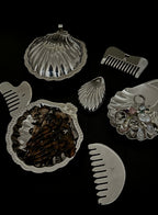A set of silver-toned shell-shaped hair accessories and hair clips arranged on a black background, including a large clamshell dish with brown hair inside, a smaller shell-shaped clip, a comb, and a shell-shaped jewelry organizer holding pearl-like beads.