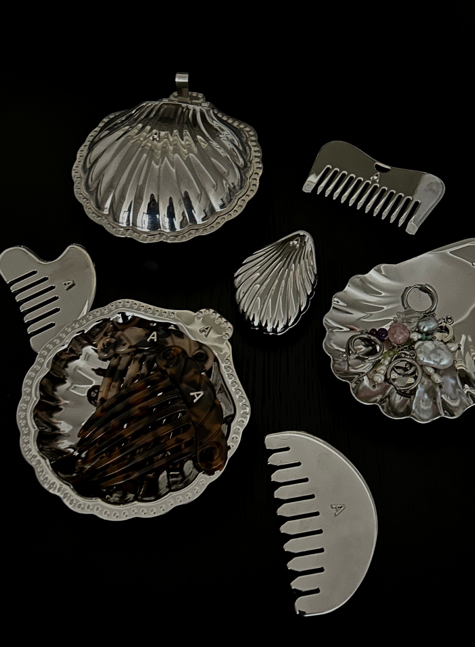 A set of silver-toned shell-shaped hair accessories and hair clips arranged on a black background, including a large clamshell dish with brown hair inside, a smaller shell-shaped clip, a comb, and a shell-shaped jewelry organizer holding pearl-like beads.