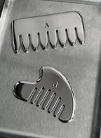 Two polished metallic grooming tools, likely ceramic or stone hair removal combs, displayed on a brushed metal surface. One comb is oriented horizontally at the top, and the other is at a diagonal angle below it.