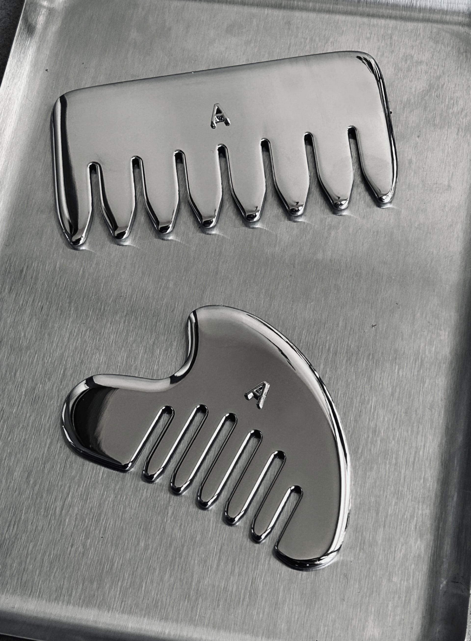 Two polished metallic grooming tools, likely ceramic or stone hair removal combs, displayed on a brushed metal surface. One comb is oriented horizontally at the top, and the other is at a diagonal angle below it.