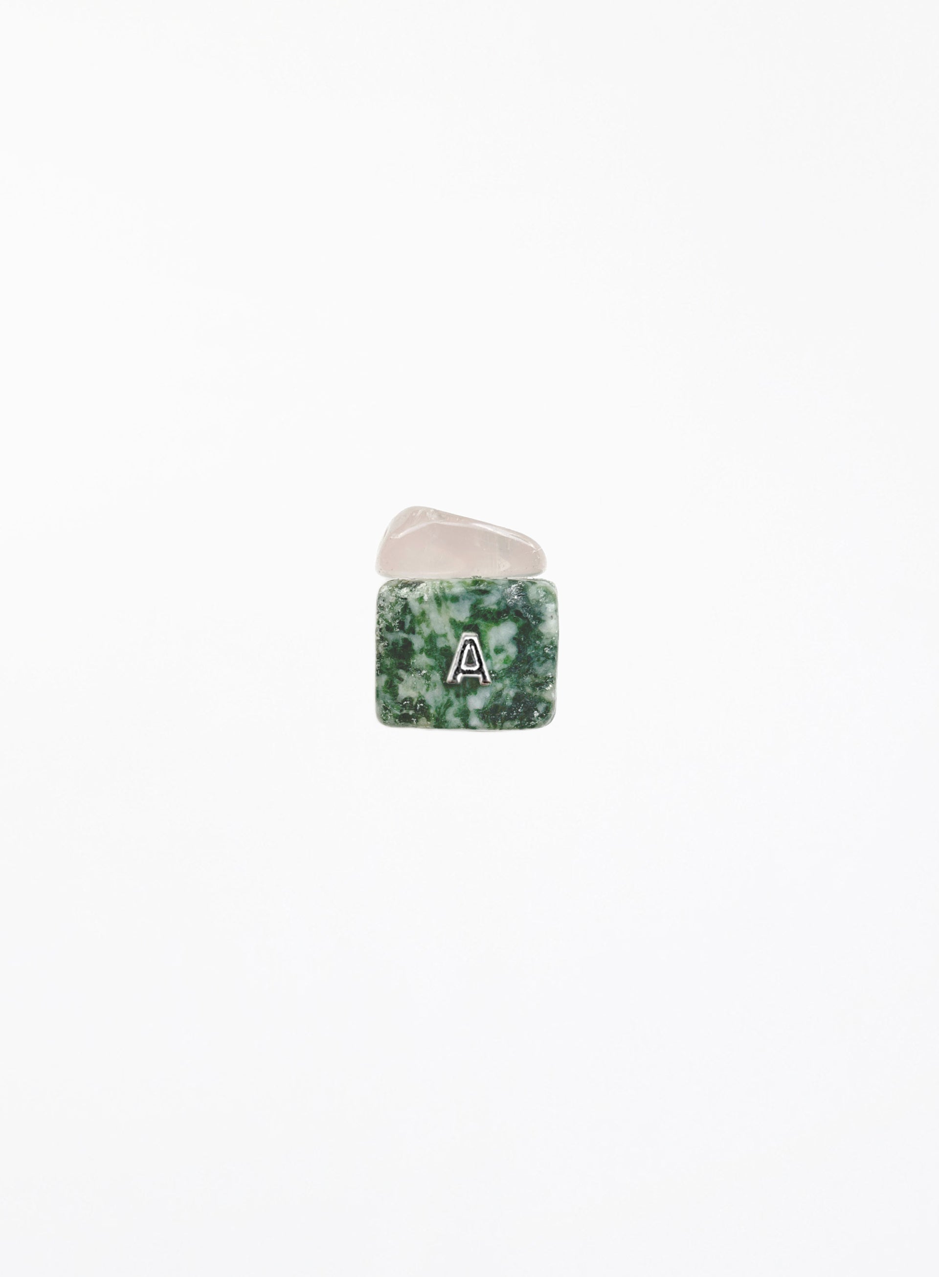 A small square gemstone charm with a green and white marbled pattern, featuring a white letter 'A' in the center, resting on a smooth, pale pink stone against a solid white background.