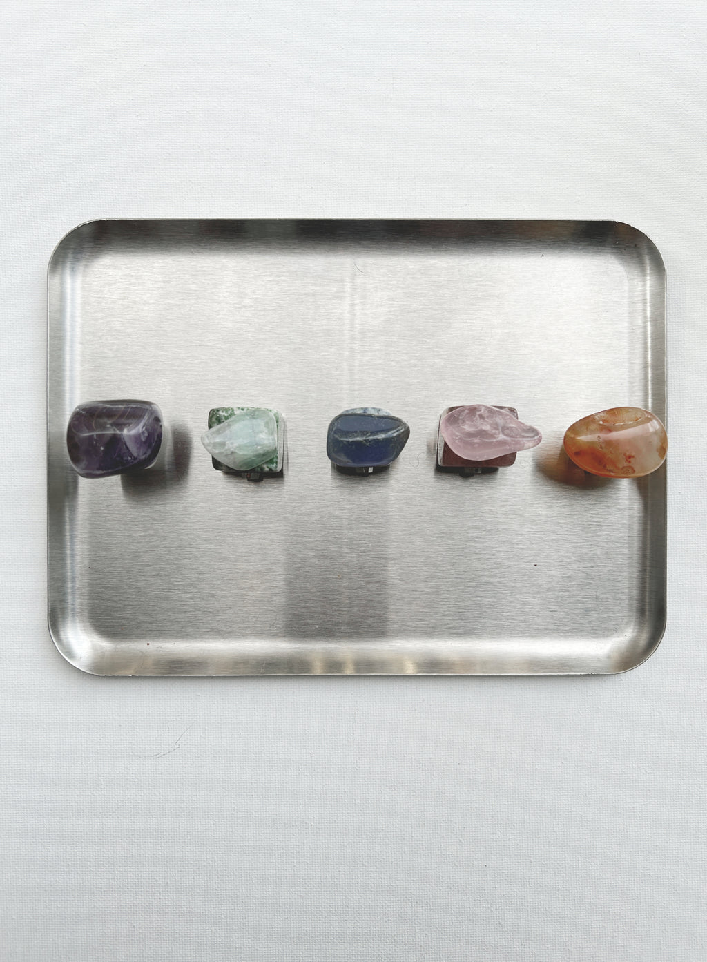 Five polished tumbled crystals in purple, green, blue, pink, and orange are lined up on a rectangular brushed metal tray against a light gray background.