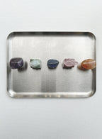 Five polished tumbled crystals in purple, green, blue, pink, and orange are lined up on a rectangular brushed metal tray against a light gray background.