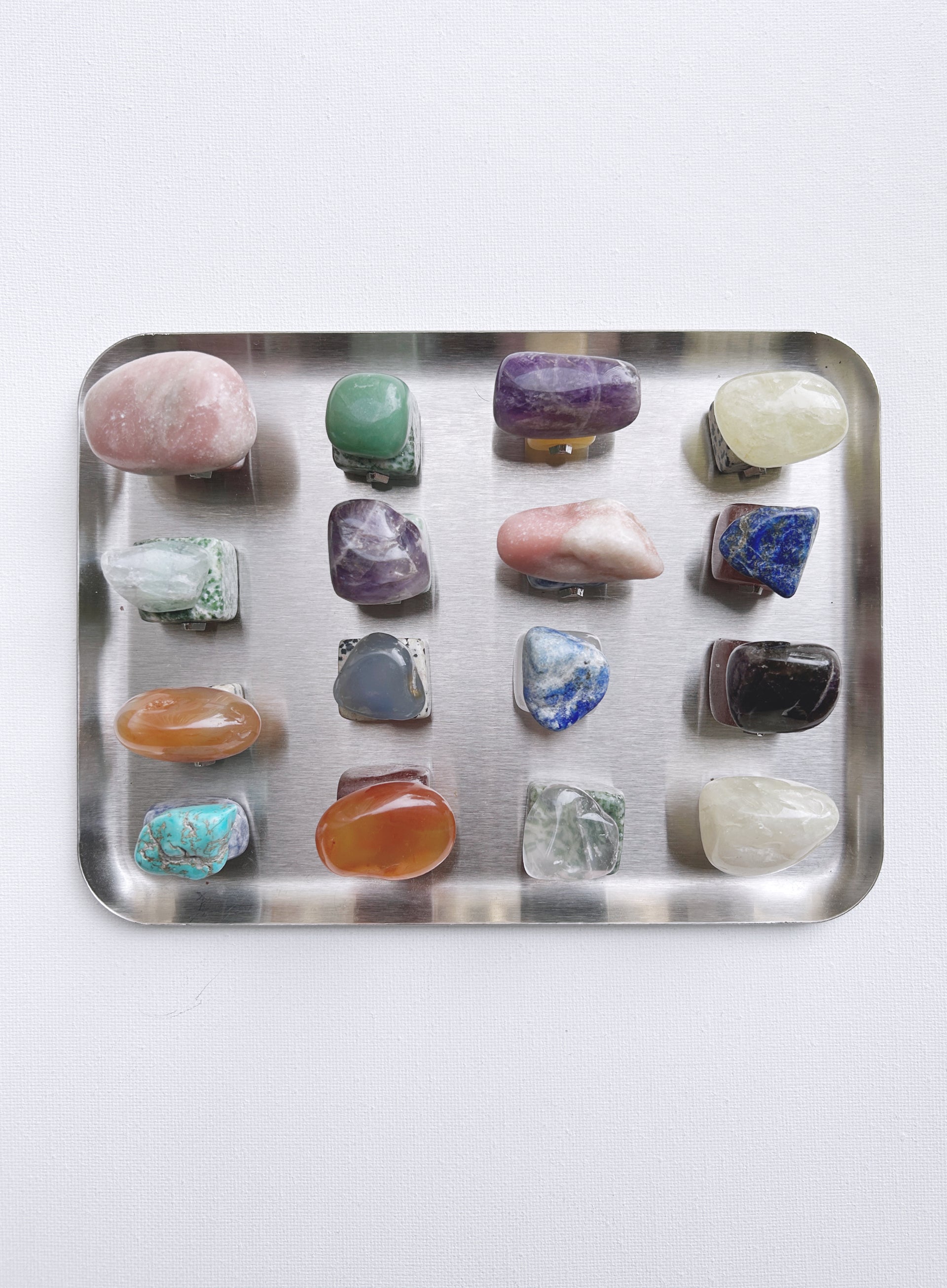 A collection of twenty different polished tumbled gemstones arranged in a grid on a rectangular silver metal tray, shot from a top-down view against a plain white background.