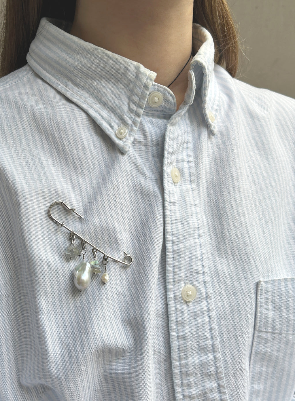 A silver safety pin brooch with dangling pearl-like beads is pinned onto a light blue and white striped button-down shirt worn by a model.