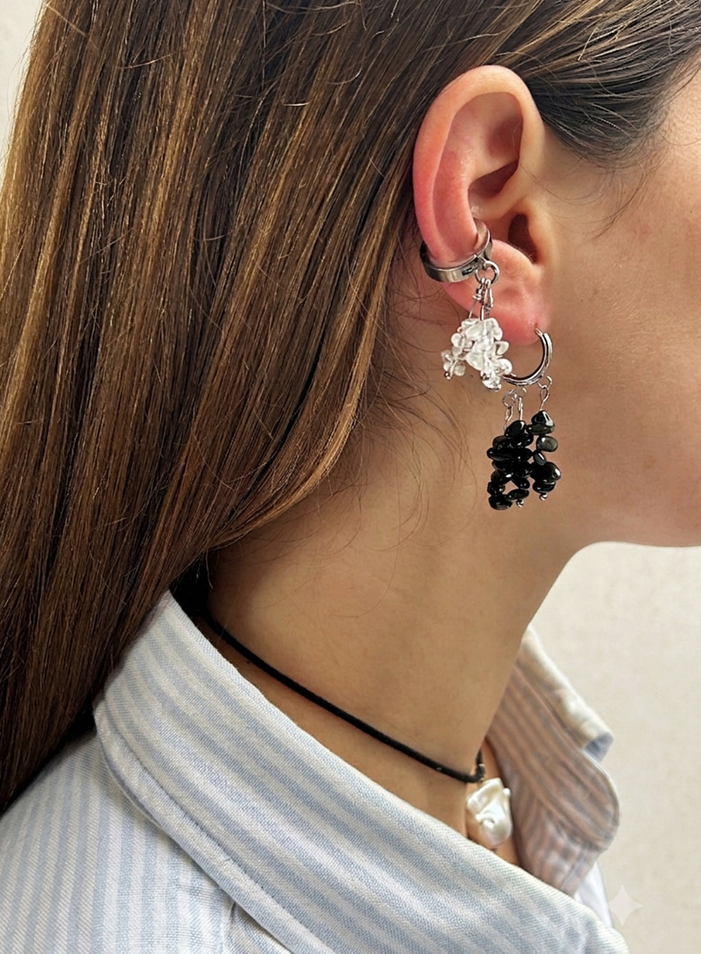 A close-up side profile of a woman wearing a silver huggie hoop earring with dangling floral charms, one in clear crystal and one in black seed beads, paired with a light blue and white striped shirt.