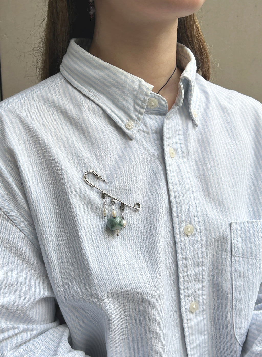 A close-up of a person wearing a white and light blue vertically striped button-down shirt, featuring a silver safety pin brooch with dangling green and white beads pinned to the chest pocket.