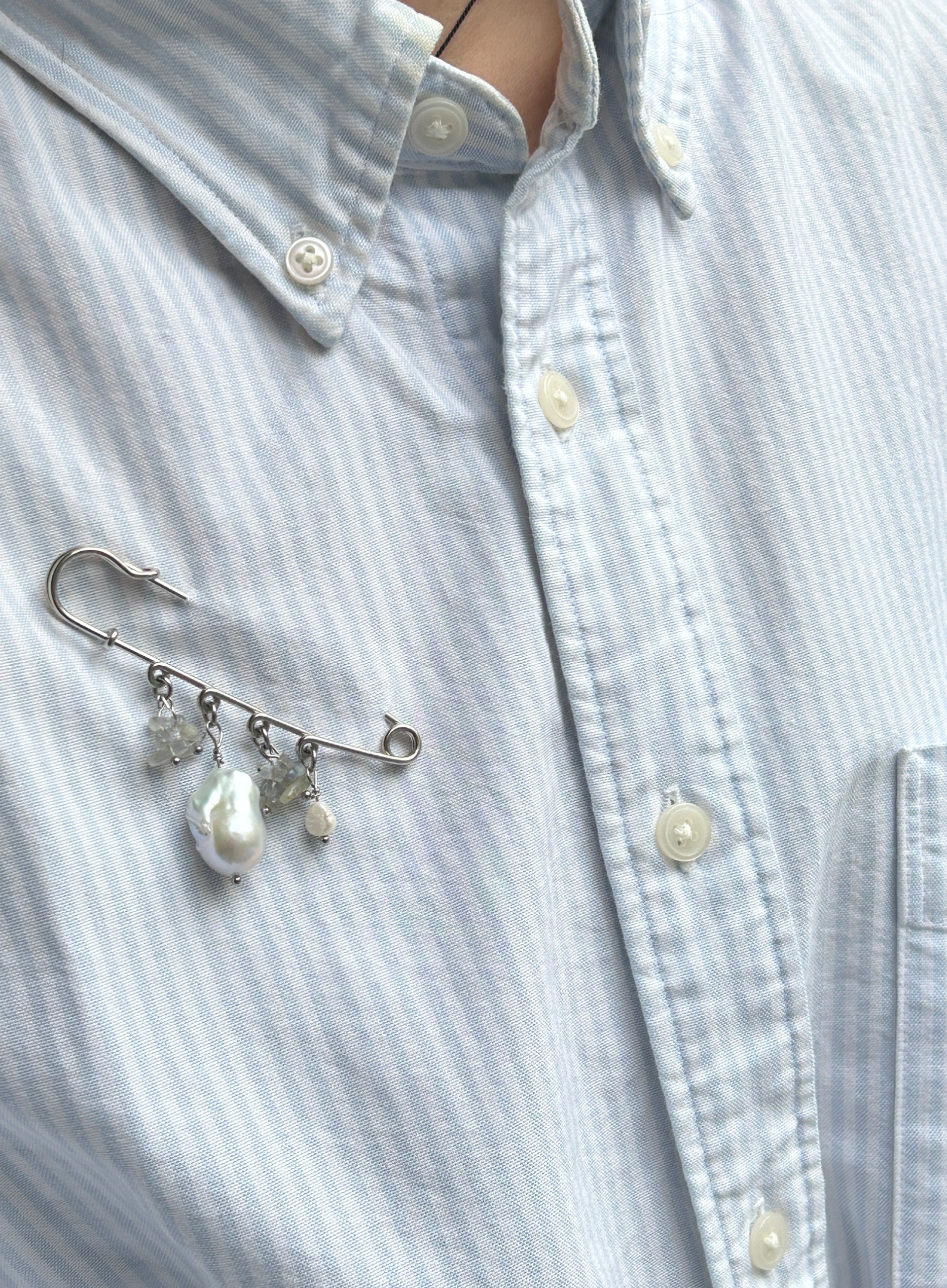 A silver safety pin brooch with dangling baroque pearls and clear crystal accents, pinned to a light blue and white striped button-down shirt.