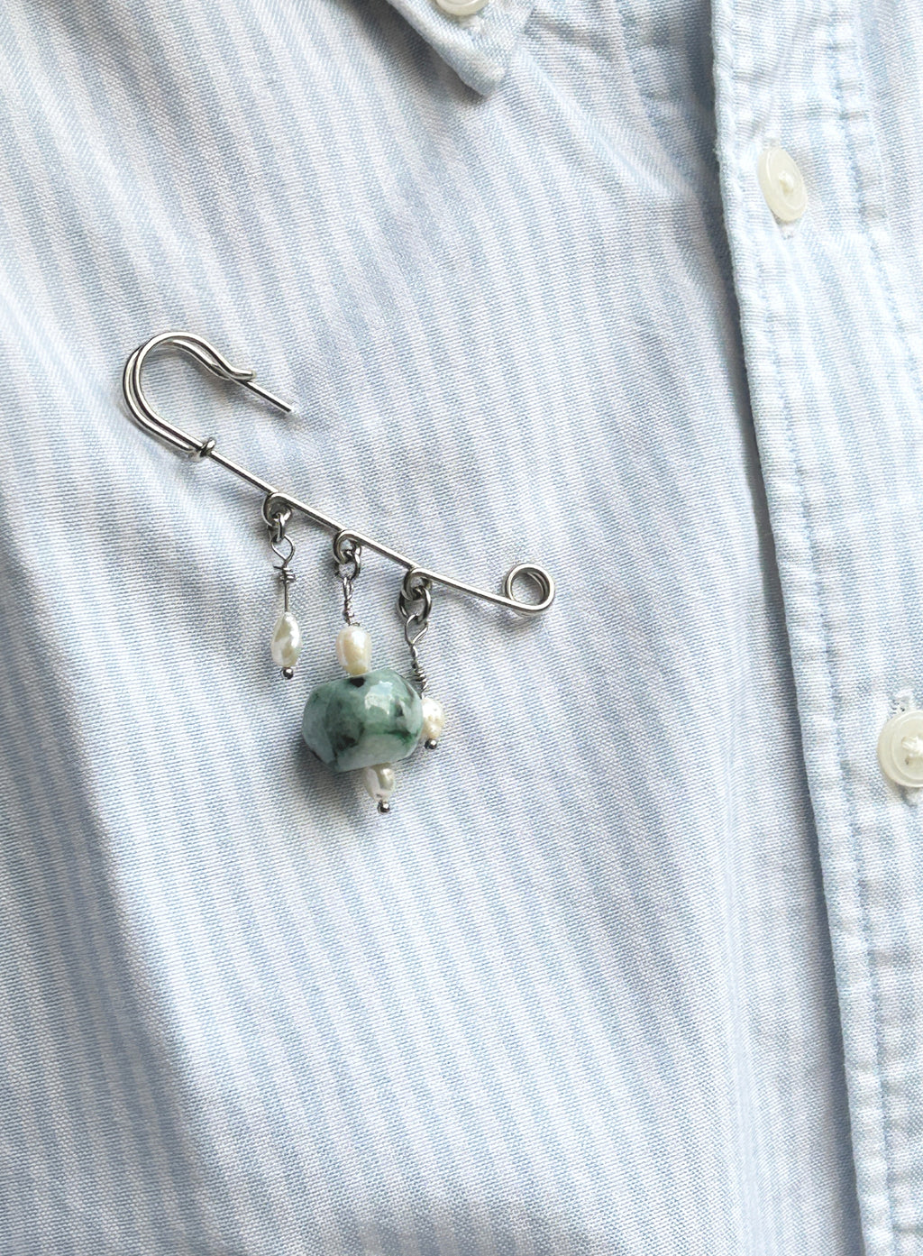 A silver safety pin brooch with dangling white pearl-like beads and a green jade bead, pinned to a light blue and white striped button-down shirt.