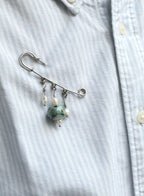 A silver safety pin brooch with dangling white pearl-like beads and a green jade bead, pinned to a light blue and white striped button-down shirt.