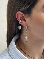A close-up of a person's ear wearing two dangling earrings: a silver-toned ear cuff with a clear faceted bead and a silver hoop earring with a silver ring, a white pearl bead, a lavender purple flower-shaped bead, and a small purple bead at the bottom.