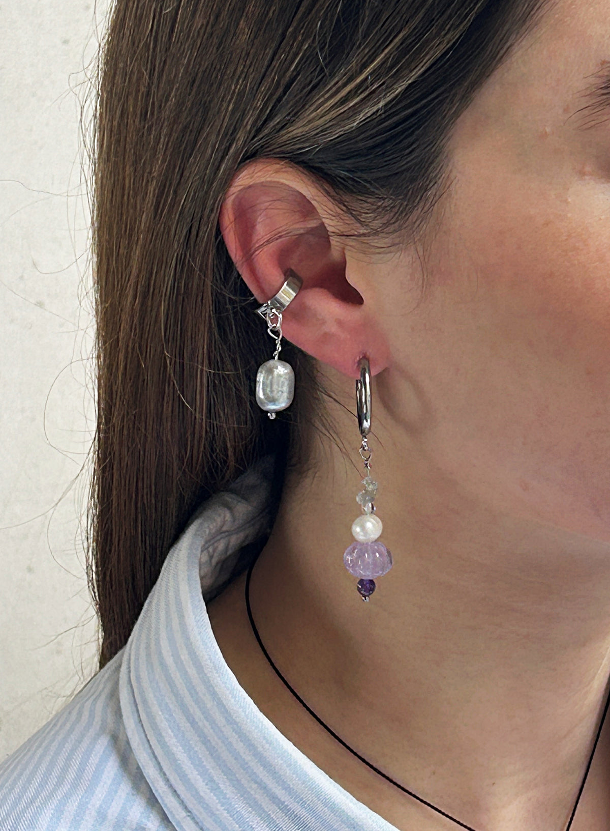 A close-up of a person's ear wearing two dangling earrings: a silver-toned ear cuff with a clear faceted bead and a silver hoop earring with a silver ring, a white pearl bead, a lavender purple flower-shaped bead, and a small purple bead at the bottom.