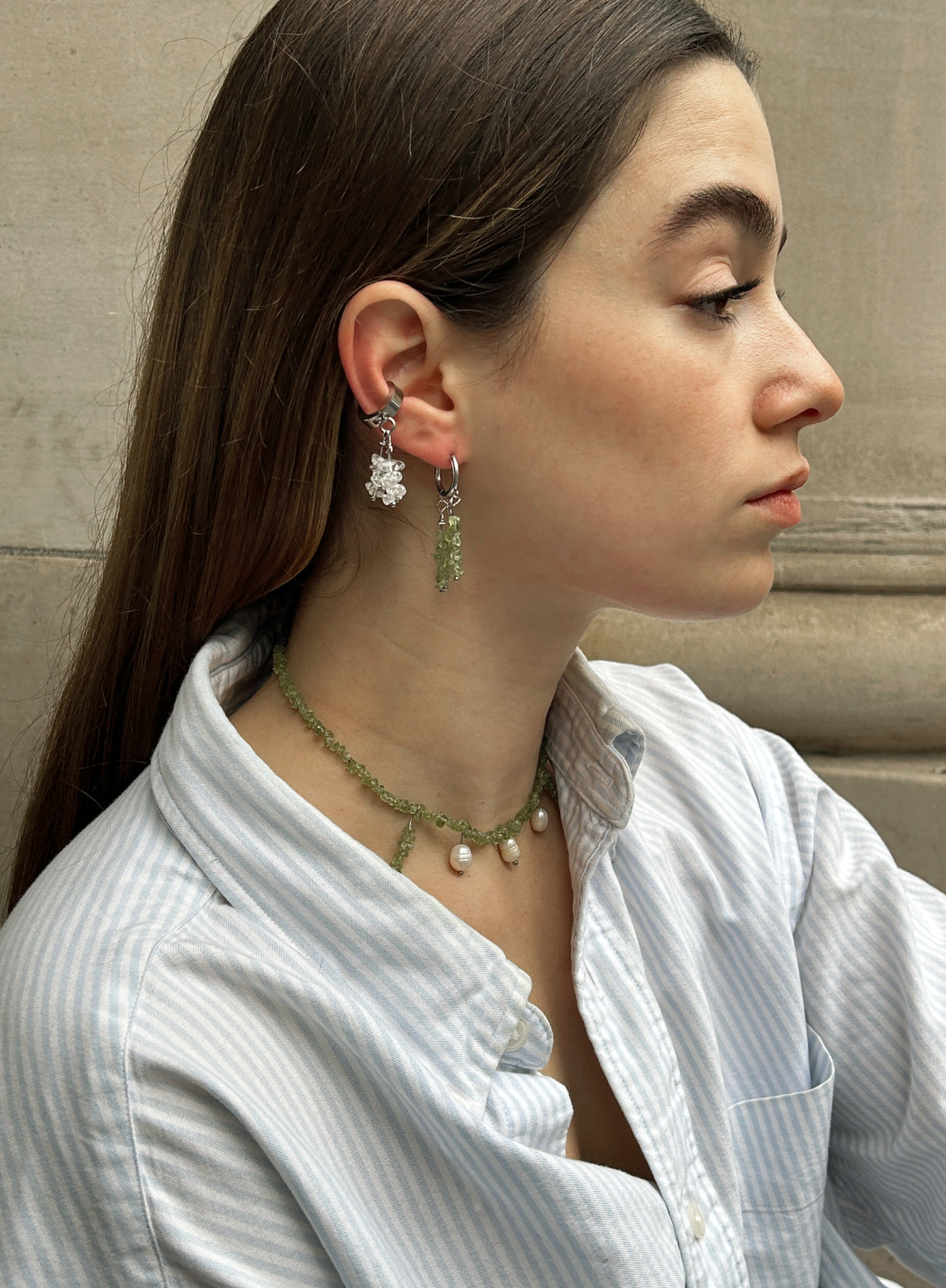 A woman in profile wearing a delicate green beaded necklace and matching drop earrings with white pearl-like accents, paired with a light blue and white striped button-up shirt.