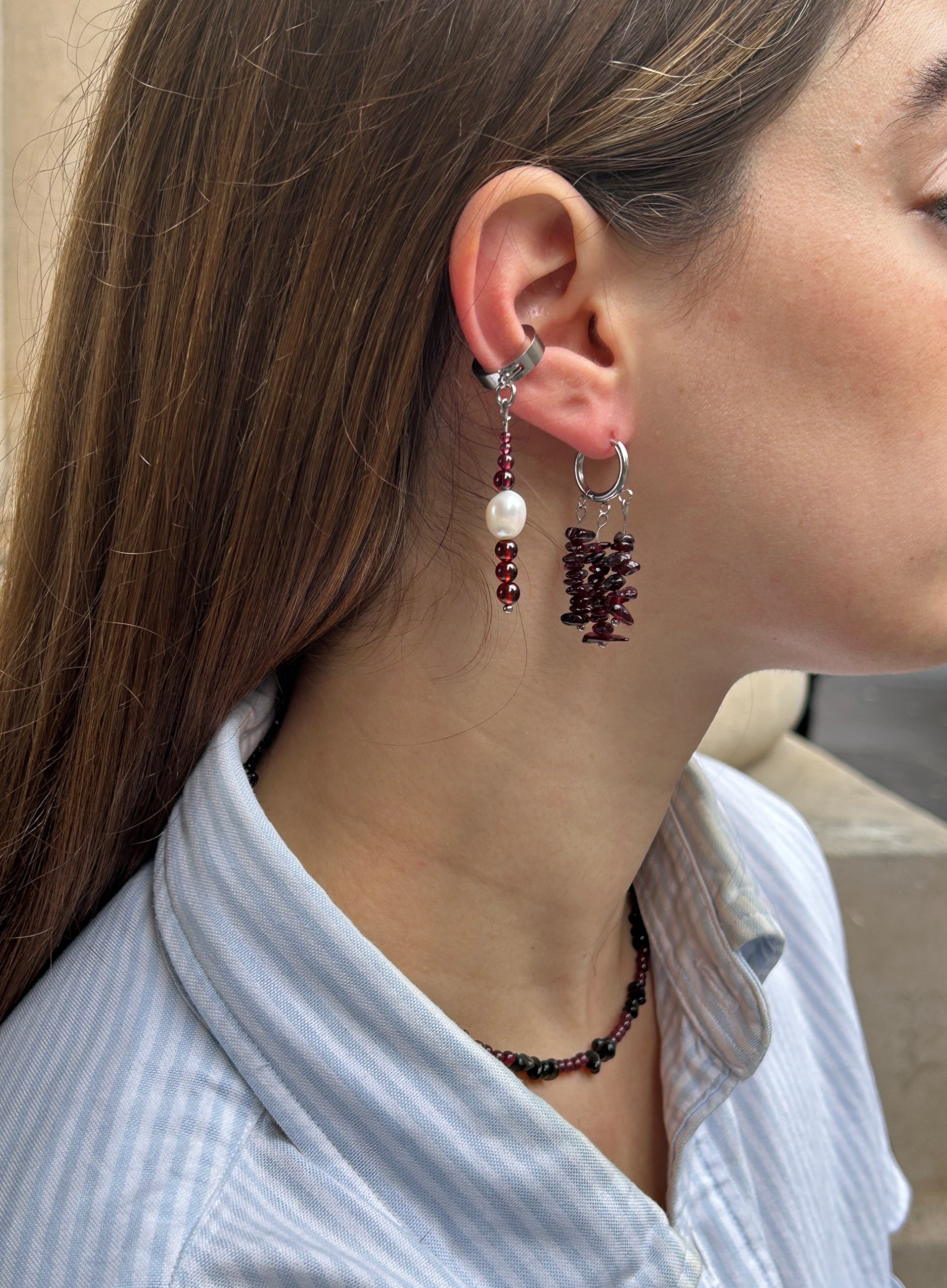 A close-up side profile of a woman wearing a silver huggie hoop earring with a dangling pearl and dark red beads, alongside another silver hoop earring with a dark red textured charm.