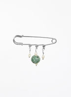 A silver safety pin brooch with three dangling charms: a large green jade bead, a small cream pearl, and a tiny silver bead.