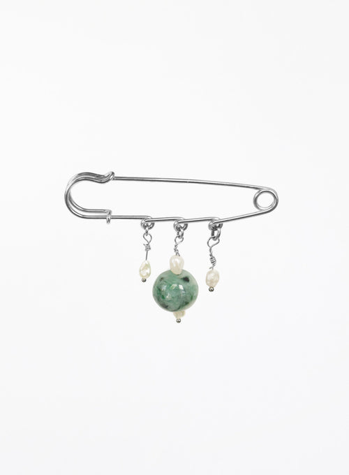 A silver safety pin brooch with three dangling charms: a large green jade bead, a small cream pearl, and a tiny silver bead.