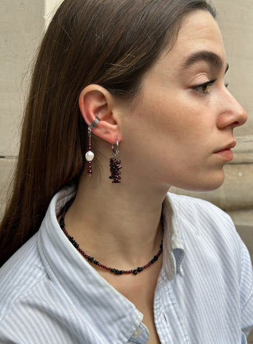 A close-up side profile of a woman wearing a silver beaded drop earring with a white pearl and dark red beads, along with a matching beaded necklace.