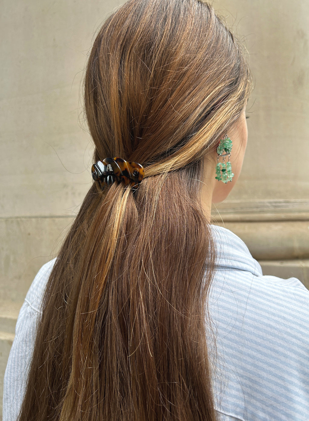 Person with long brown hair tied back with a hair tie, wearing green earrings against a neutral background