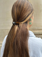 Person with long brown hair tied back with a hair tie, wearing green earrings against a neutral background