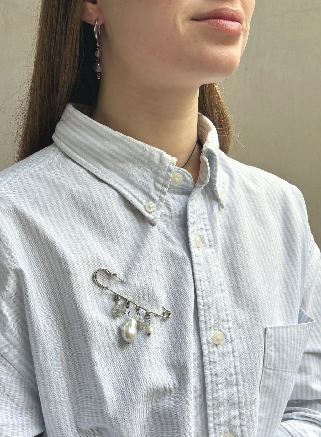 A close-up of a person wearing a light blue and white striped button-down shirt, featuring a silver safety-pin style brooch with dangling white faux pearls pinned to the chest pocket.