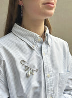 A close-up of a person wearing a light blue and white striped button-down shirt, featuring a silver safety-pin style brooch with dangling white faux pearls pinned to the chest pocket.