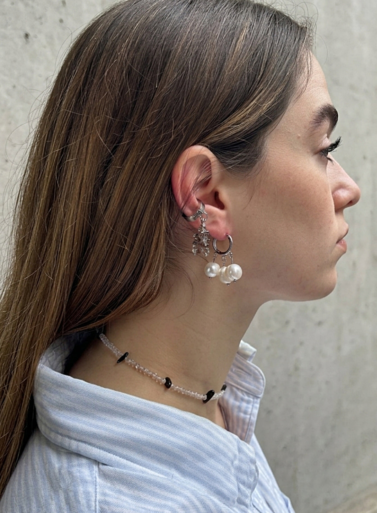 A close-up side profile of a woman wearing silver hoop earrings with white pearl drops and a beaded choker necklace, paired with a light blue striped shirt against a textured gray wall.