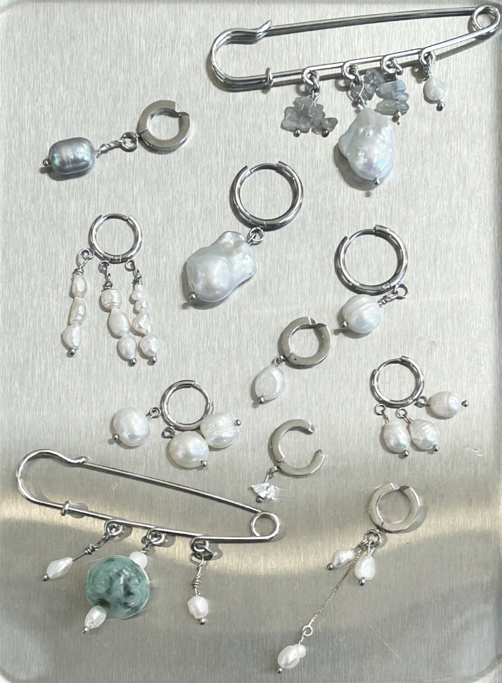 A collection of silver-toned body jewelry and earrings displayed on a brushed metal surface, including safety pins, hoop earrings, dangling pearl bead drops, and irregular pearl charms.