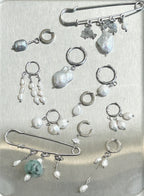 A collection of silver-toned body jewelry and earrings displayed on a brushed metal surface, including safety pins, hoop earrings, dangling pearl bead drops, and irregular pearl charms.