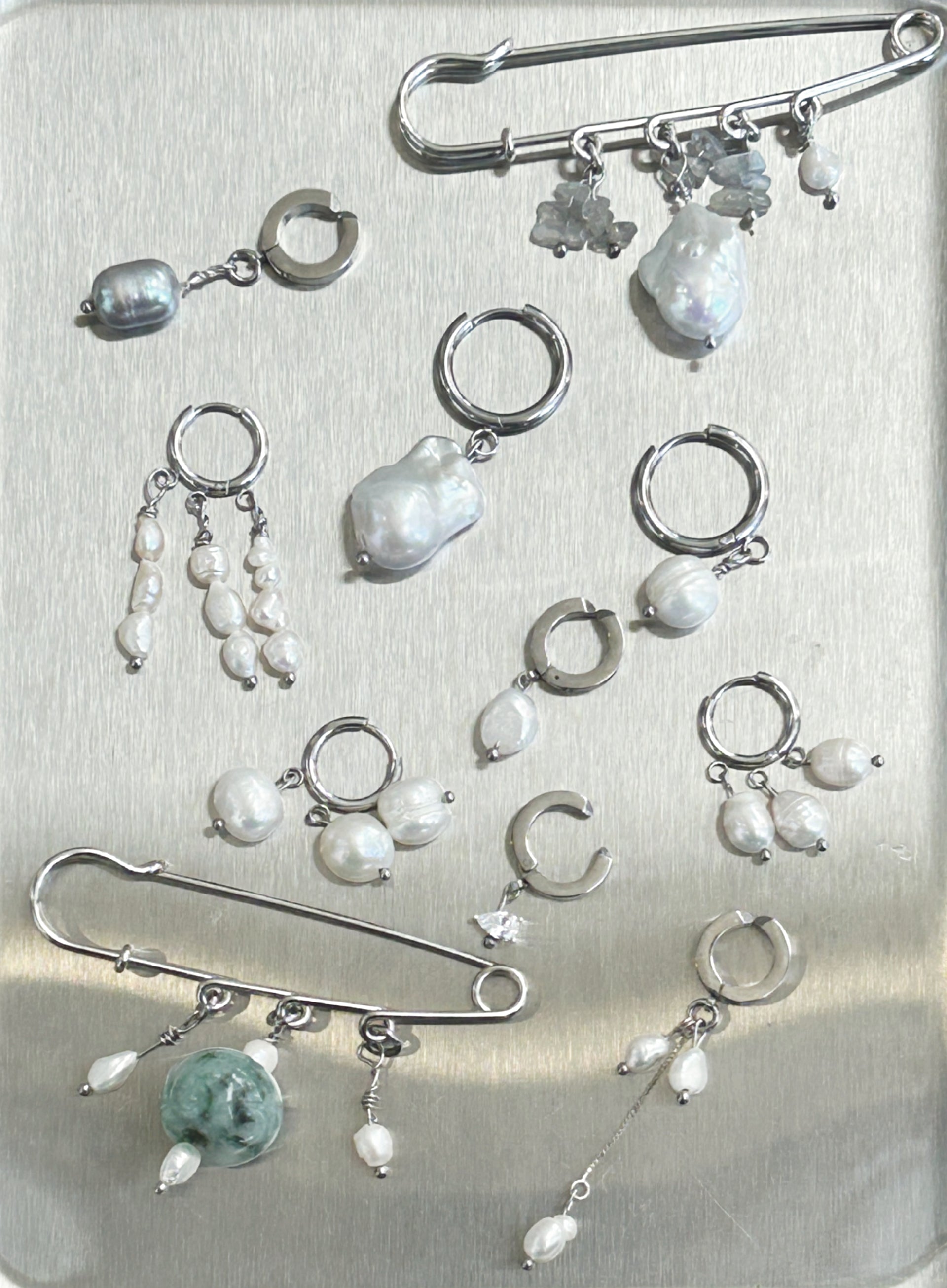 A collection of silver-toned body jewelry and earrings displayed on a brushed metal surface, including safety pins, hoop earrings, dangling pearl bead drops, and irregular pearl charms.