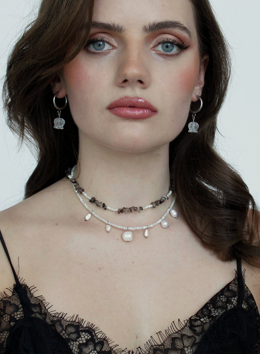 A close-up of a woman wearing layered necklaces and matching dangling earrings. The jewelry includes a dark beaded choker, a white pearl bead choker, and silver hoop earrings with clear crystal pendants.