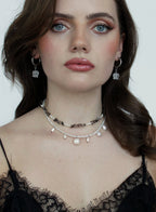 A close-up of a woman wearing layered necklaces and matching dangling earrings. The jewelry includes a dark beaded choker, a white pearl bead choker, and silver hoop earrings with clear crystal pendants.