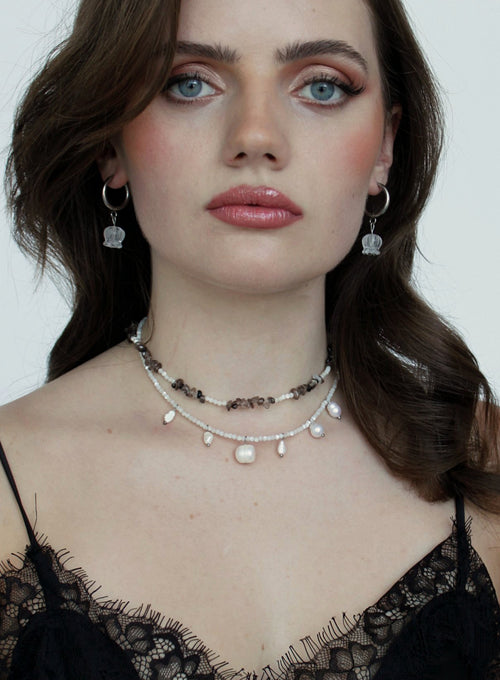 A close-up of a woman wearing layered necklaces and matching dangling earrings. The jewelry includes a dark beaded choker, a white pearl bead choker, and silver hoop earrings with clear crystal pendants.