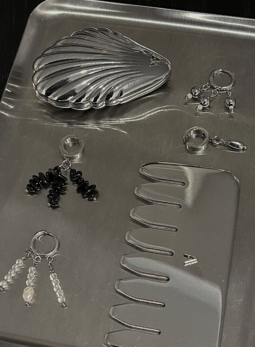 A silver jewelry organizer tray containing a shell-shaped coin purse, several pairs of dangling earrings, a small comb, and a rectangular case, photographed on a dark background.