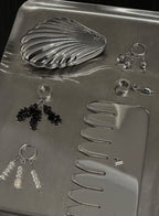 A silver jewelry organizer tray containing a shell-shaped coin purse, several pairs of dangling earrings, a small comb, and a rectangular case, photographed on a dark background.