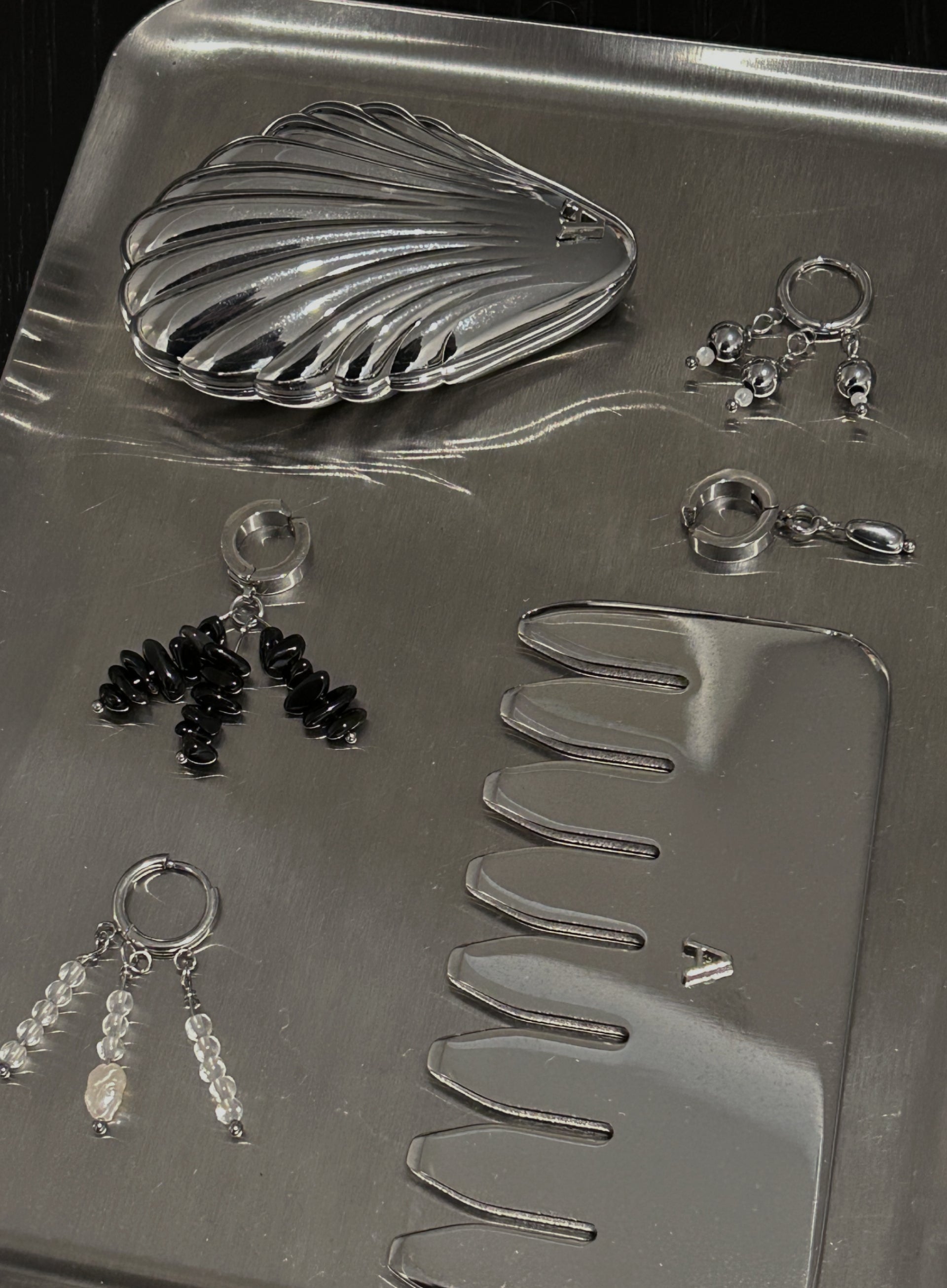 A silver jewelry organizer tray containing a shell-shaped coin purse, several pairs of dangling earrings, a small comb, and a rectangular case, photographed on a dark background.
