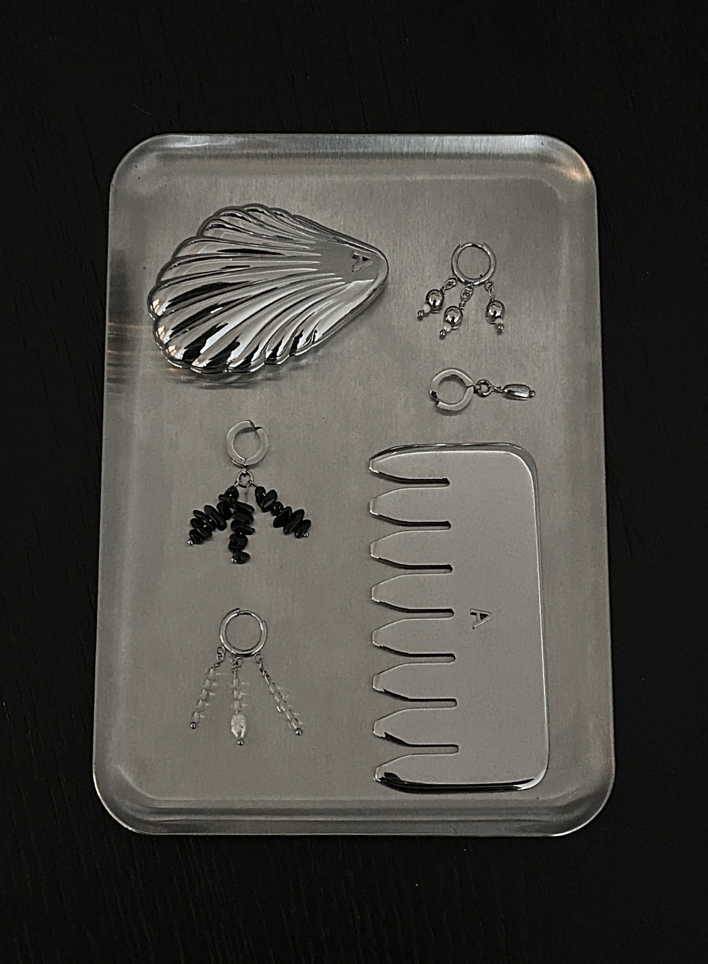 A silver metal jewelry organizer tray containing a clam shell-shaped mirror, a flat comb, several small hoop and stud earrings, and a clasp, photographed on a dark background.