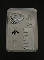 A silver metal jewelry organizer tray containing a clam shell-shaped mirror, a flat comb, several small hoop and stud earrings, and a clasp, photographed on a dark background.
