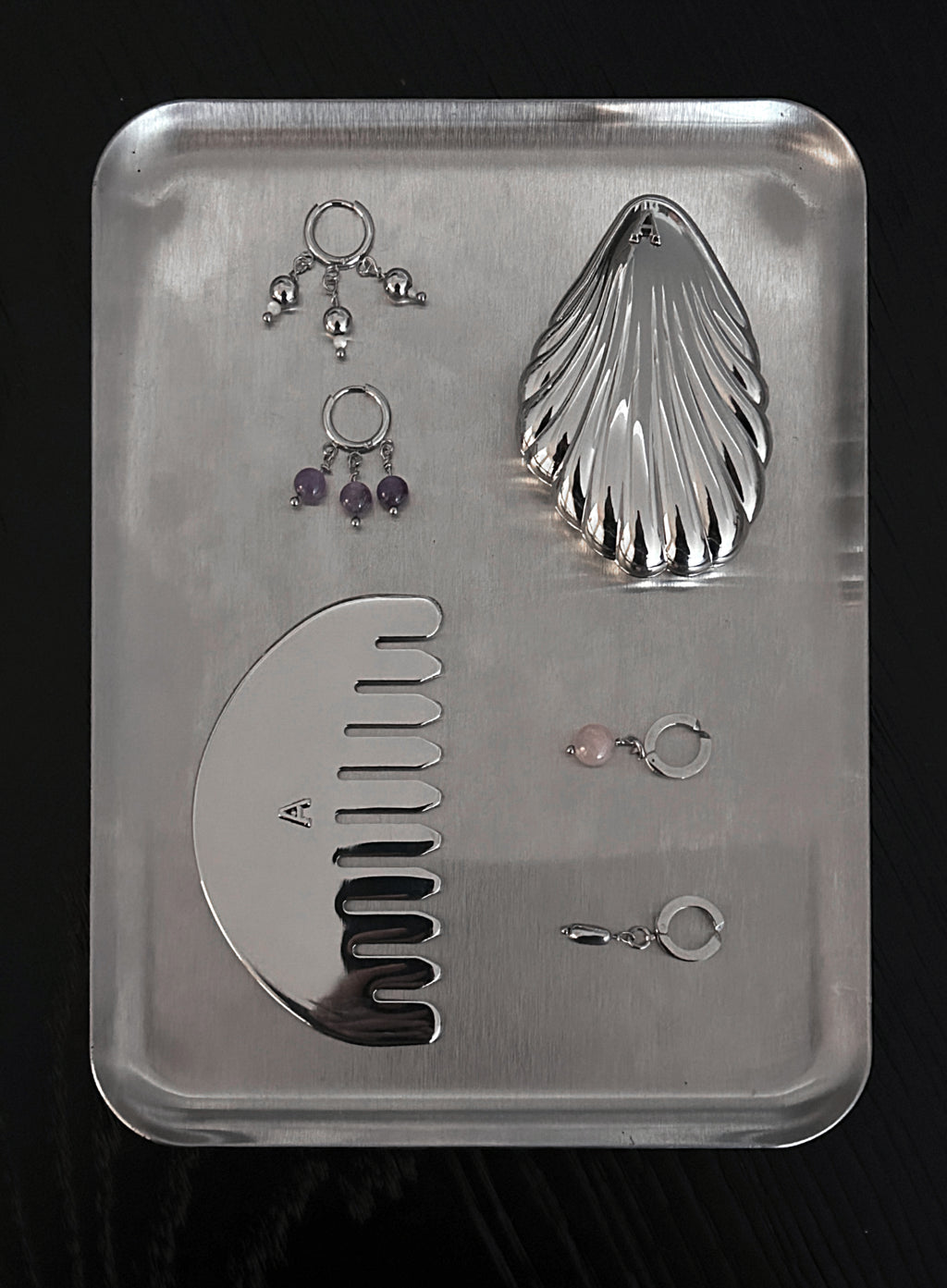 A set of silver-toned body jewelry and a large silver comb arranged on a rectangular metal tray against a black background. The set includes multiple ear cuffs, two pairs of dangling earrings with purple and pink stones, and a curved comb.