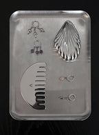 A set of silver-toned body jewelry and a large silver comb arranged on a rectangular metal tray against a black background. The set includes multiple ear cuffs, two pairs of dangling earrings with purple and pink stones, and a curved comb.