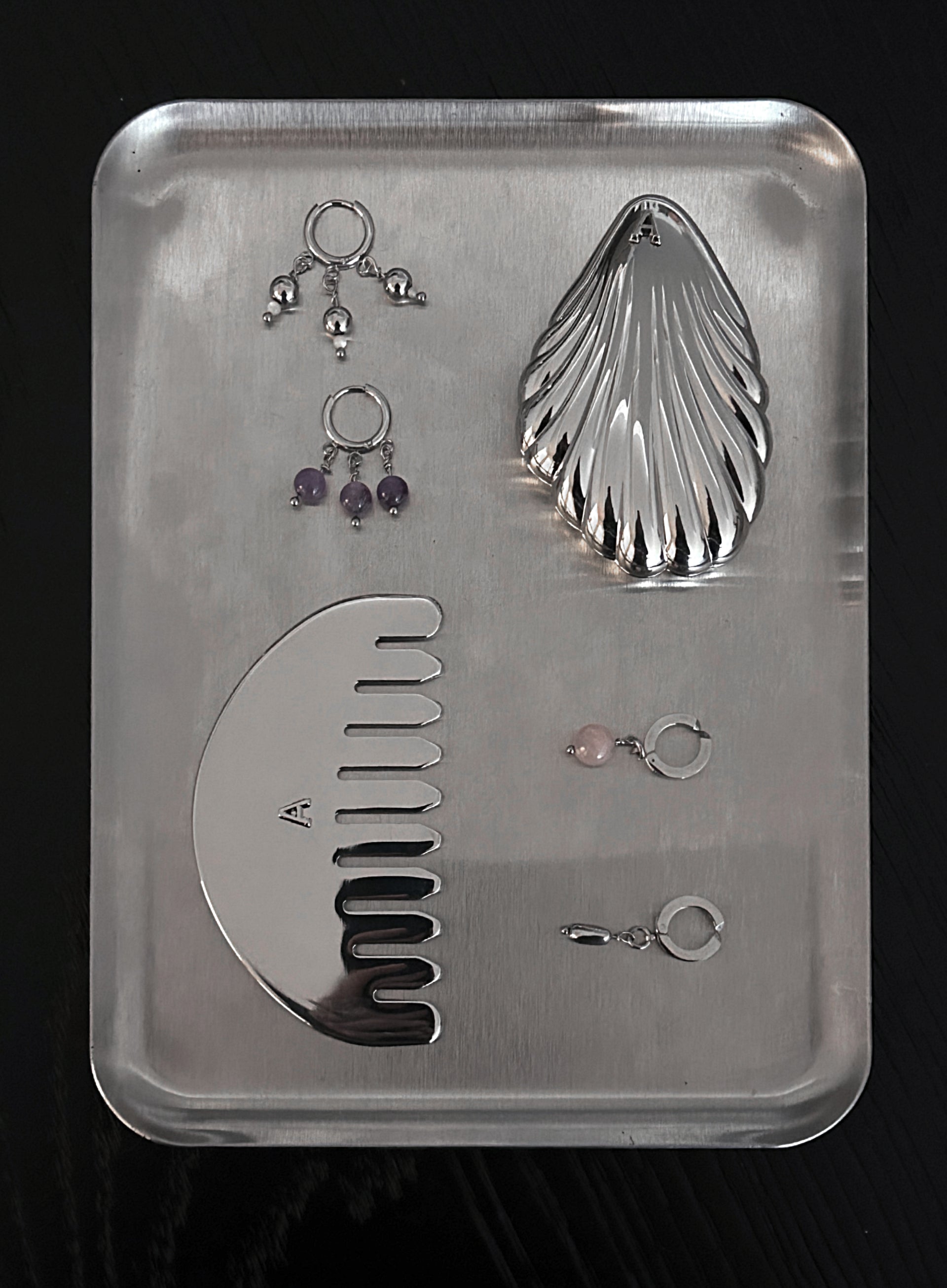 A set of silver-toned body jewelry and a large silver comb arranged on a rectangular metal tray against a black background. The set includes multiple ear cuffs, two pairs of dangling earrings with purple and pink stones, and a curved comb.