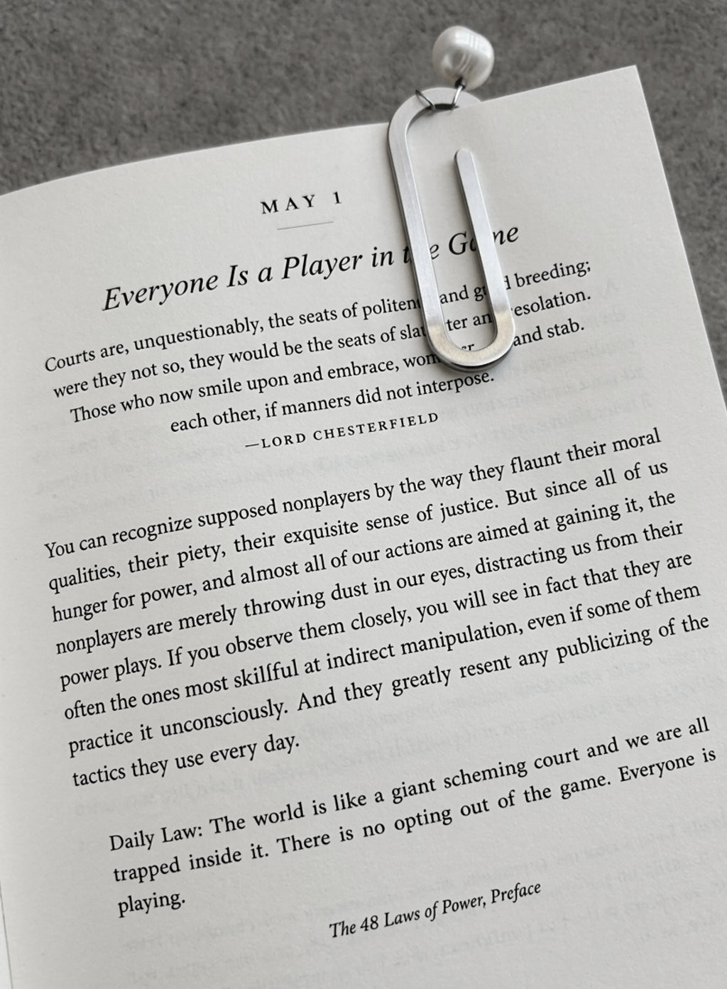 A steel paper-clip bookmark clipped across the top of a book, photographed on a textured gray surface.