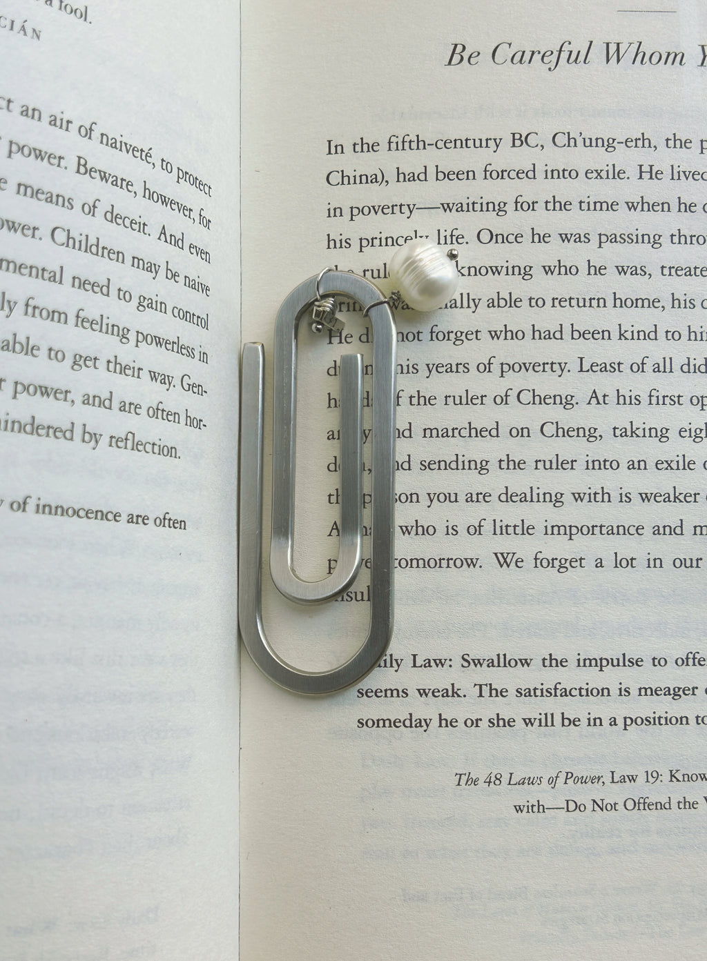 A silver-toned metal paperclip-shaped bookmark with a small white pearl accent and inital charm, resting on an open book page.