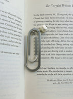A silver-toned metal paperclip-shaped bookmark with a small white pearl accent and inital charm, resting on an open book page.